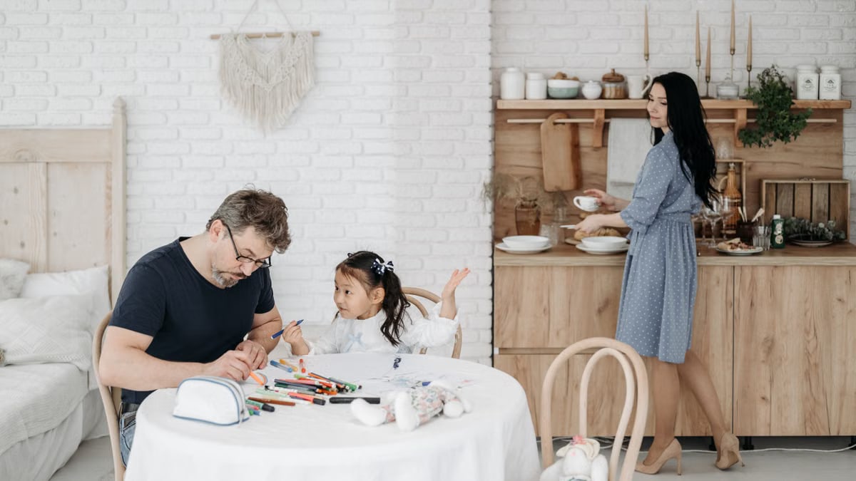 Father and daughter bond over crafts while mother prepares food in a cozy home setting.